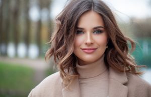 A woman with wavy brown hair and blue eyes is wearing a beige turtleneck and coat, standing outdoors with a blurred background of trees and greenery.