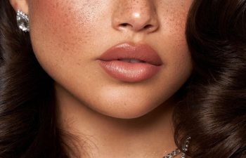 Close-up of a womans face showing smooth skin with freckles, full matte pink lips, wavy dark brown hair, and part of a diamond earring and necklace visible.