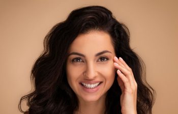 A woman with long, wavy dark hair smiles at the camera, her hand gently touching her face. She has clear skin and is photographed against a soft beige background.