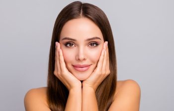 A woman with straight brown hair touches her face with both hands, smiling softly. She has clear skin, natural makeup, and looks directly at the camera against a plain light gray background.