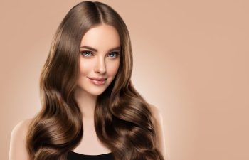 A woman with long, shiny, wavy brown hair and blue eyes smiles softly against a light beige background. She is wearing a black top.